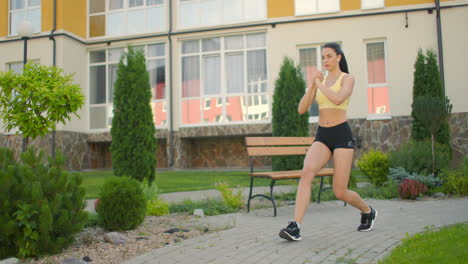 A-woman-makes-lunges-on-a-sidewalk-path-in-a-park-in-an-urban-environment.-Slow-motion-shooting-of-a-woman-in-sportswear-steps-with-a-lunge